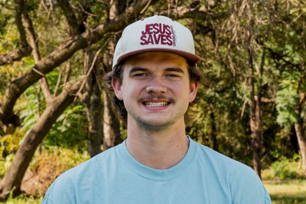 Young man wearing a light blue T-shirt and a white cap with red 'JESUS SAVES' text, outdoors in a park-like setting.