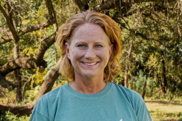 Smiling woman with shoulder-length blonde hair in a teal shirt, standing in a sunlit park with trees.