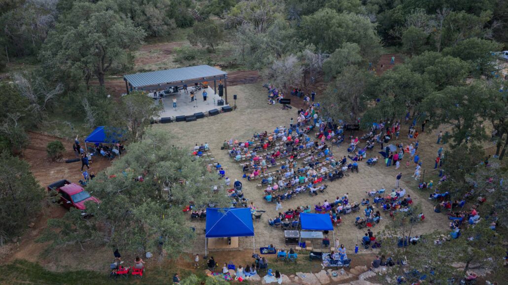 Aerial view of an outdoor concert in a forested park: a stage with musicians and a large audience seated on chairs.
