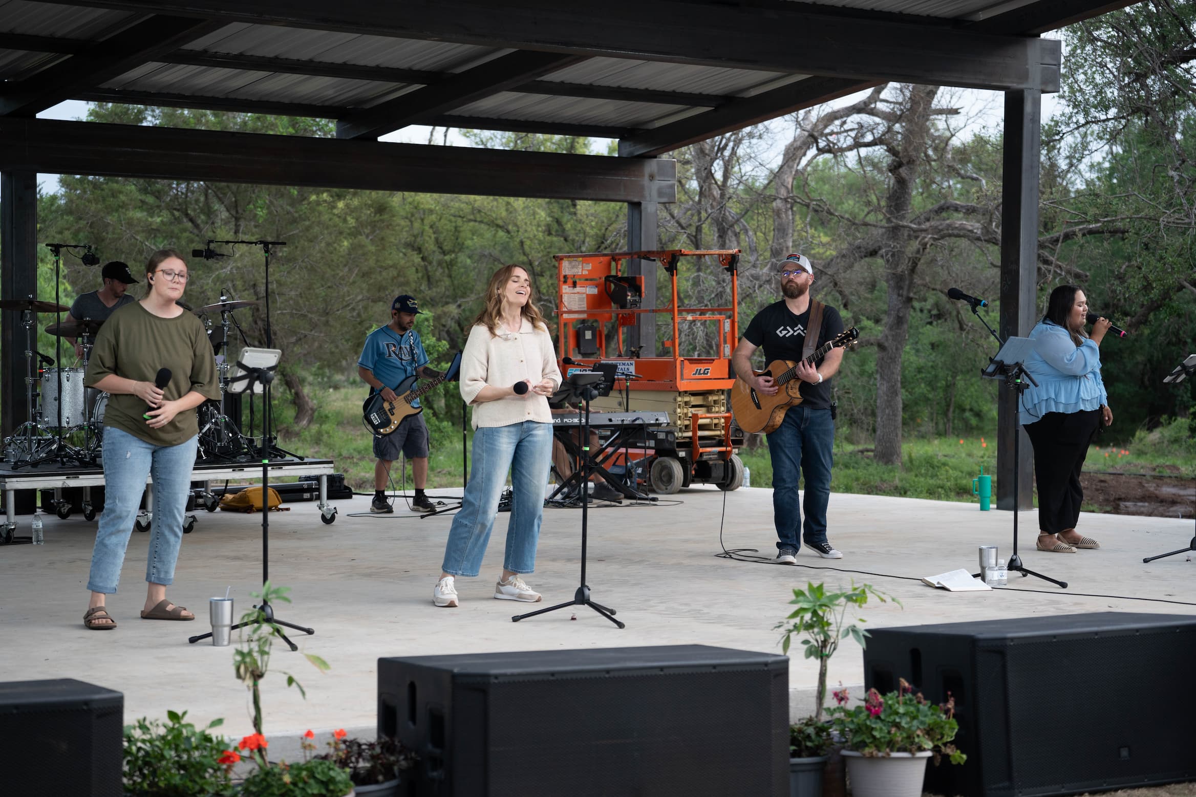 A group of musicians performing on an outdoor stage: singer center, guitarist to the right, drummer to the left, all under a shelter with trees behind.