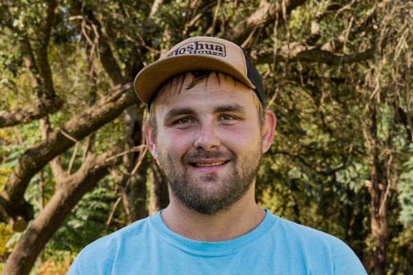 Portrait of a man in a blue shirt and tan cap, smiling with trees in the background outdoors.