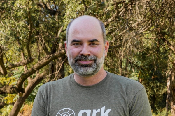 Middle-aged man with a gray beard standing outdoors among trees, wearing a green ark t-shirt and smiling at the camera.