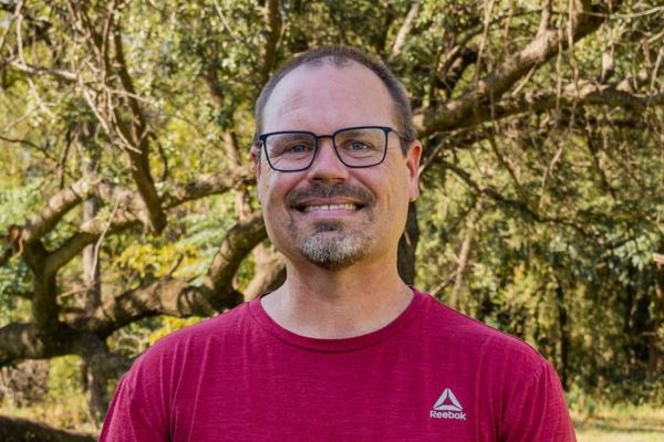 Smiling man with glasses and short beard outdoors, wearing a red Reebok t-shirt, trees in the background.