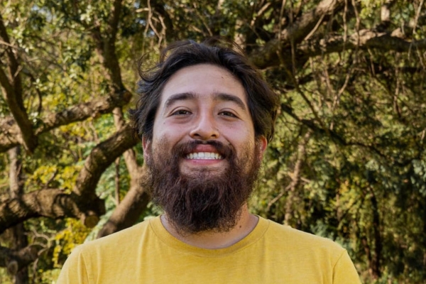Smiling man with a full beard in a yellow t-shirt standing outdoors among trees.