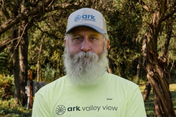 Man with gray beard wearing a lime ark Valley View shirt and gray ark cap, standing outdoors among trees.